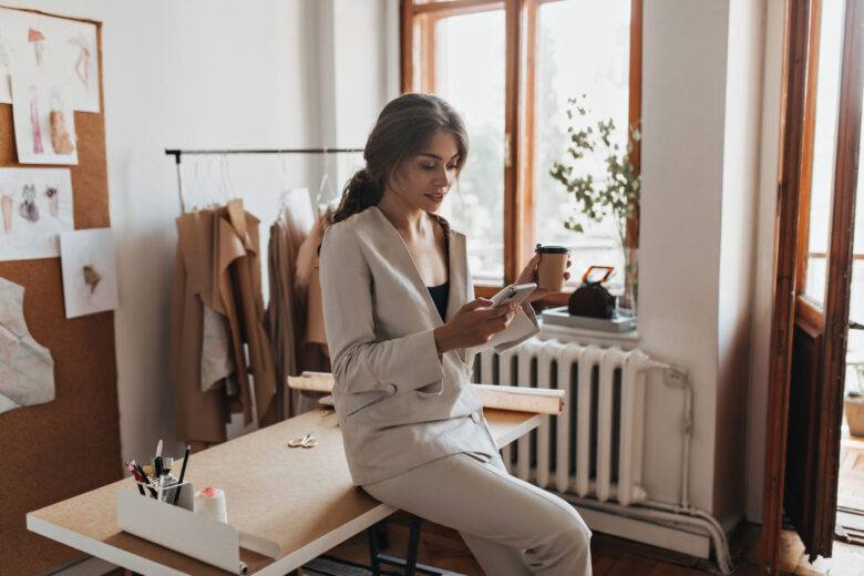 Dark haired lady in white suit holding coffee and phone. Charming woman in linen pants and beige blouse chatting in cellphone and drinking tea